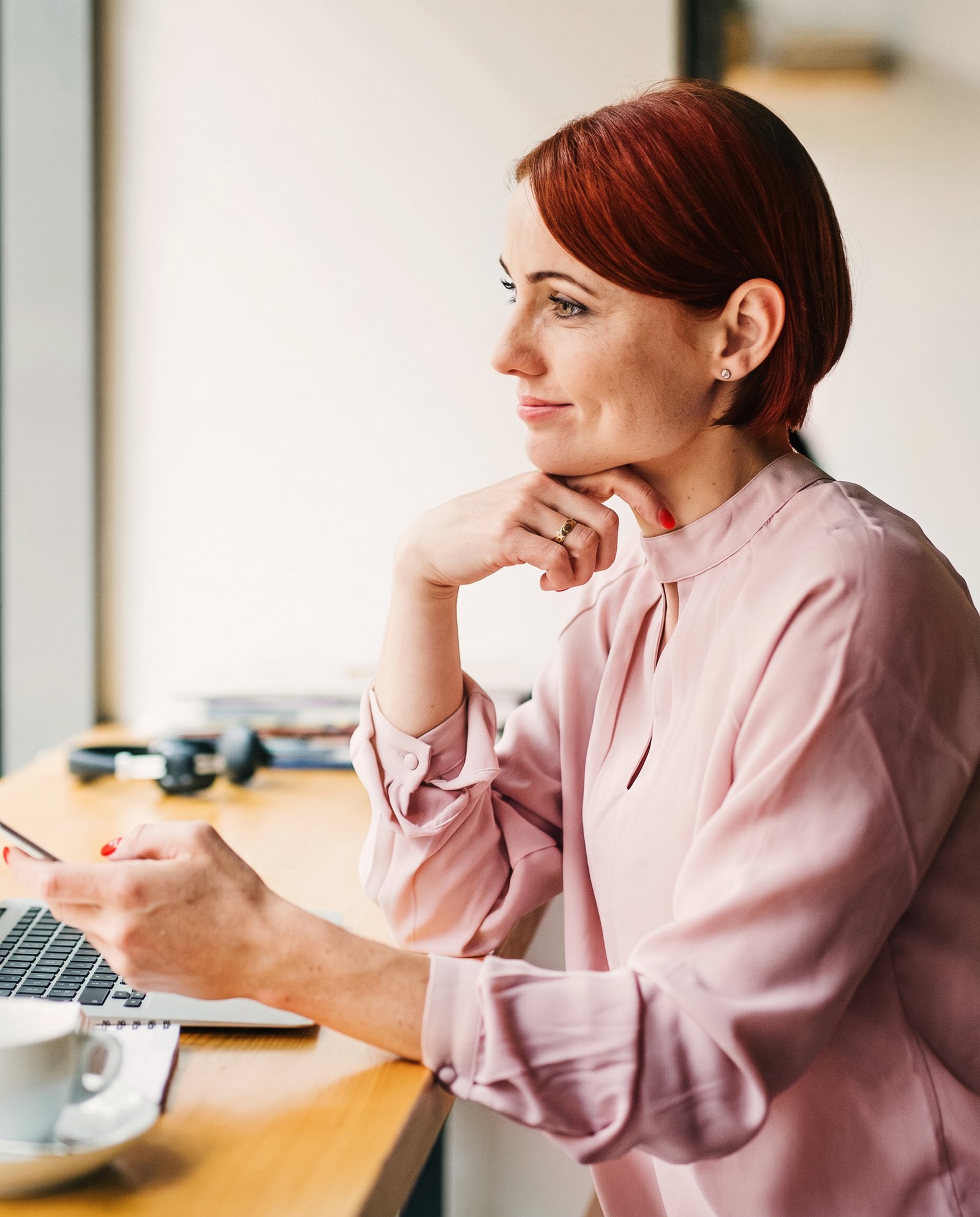 A woman with short red hair in a pale pink blouse sitting at a wooden desk, holding her phone, laptop and coffee cup in front of her